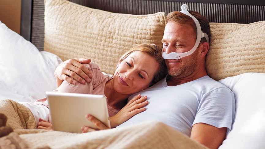 Couple lying in bed, looking at a tablet, and the man is wearing a mask for sleep apnea treatment.